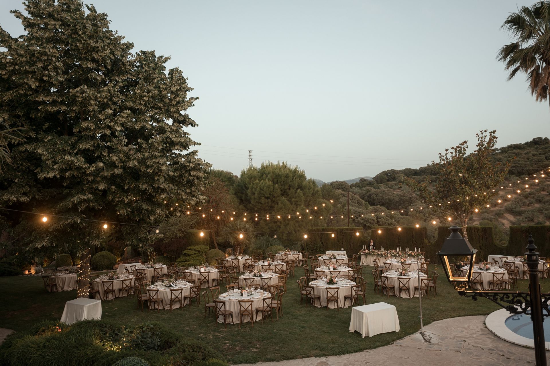 Recepción de boda nocturna al aire libre con mesas redondas, guirnaldas de luces y árboles en un entorno ajardinado.