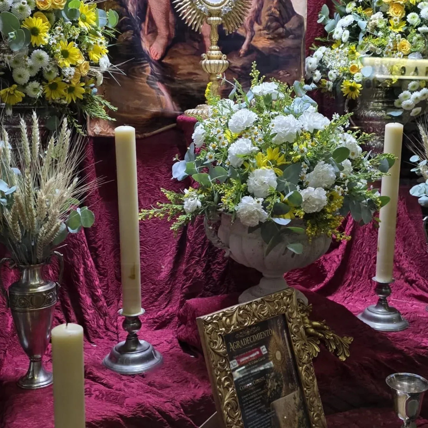 Altar conmemorativo floral con flores blancas, velas y un homenaje enmarcado sobre tela color burdeos.