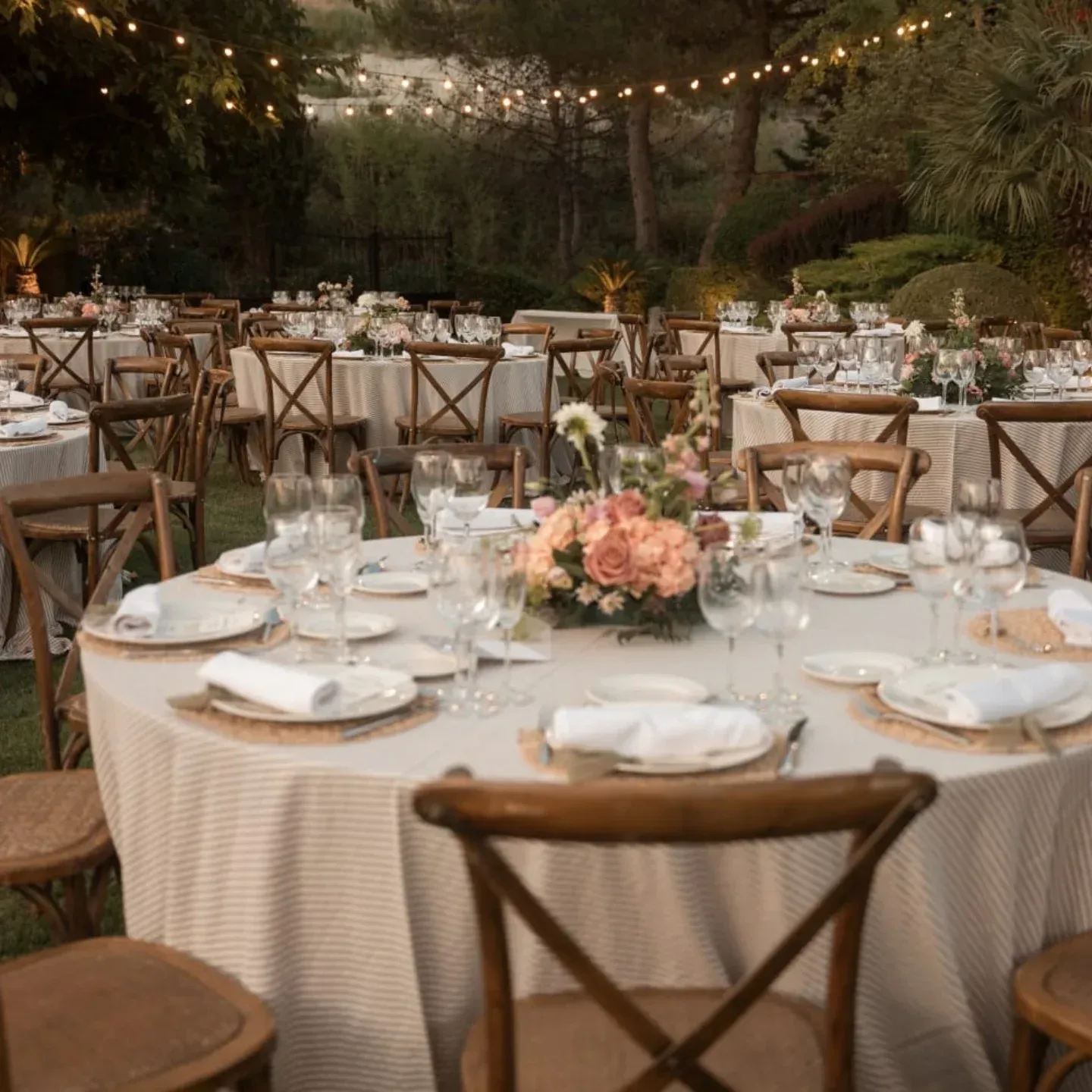 Mesas redondas para banquetes con manteles blancos y centros de mesa florales, dispuestas al aire libre bajo guirnaldas de luces al atardecer.