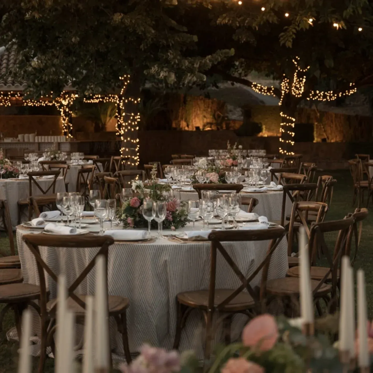Recepción con cena al aire libre con mesas redondas, sillas de madera, velas y guirnaldas de luces bajo los árboles.