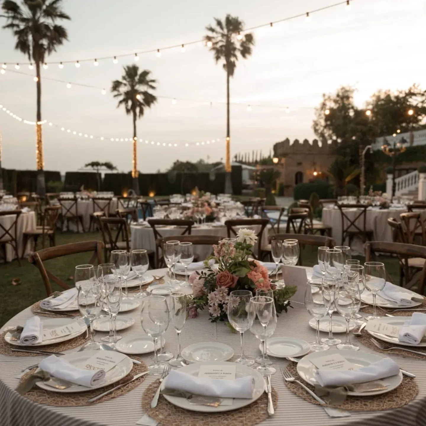 Elegante cena al aire libre con mesas redondas, guirnaldas de luces y palmeras al atardecer.