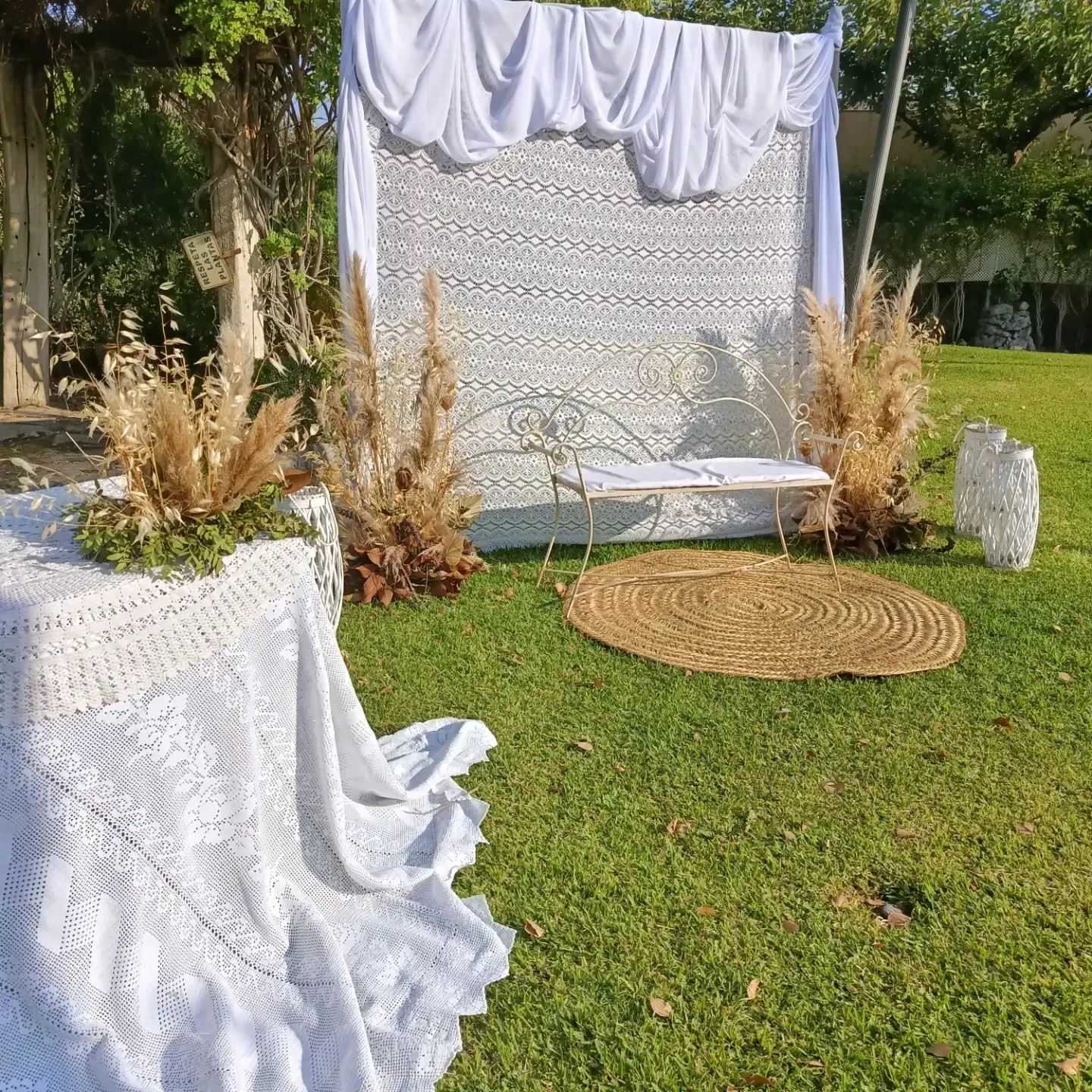 Fondo exterior cubierto con tela blanca, hierba de la pampa dorada, mesa cubierta de encaje y alfombra tejida sobre un césped.