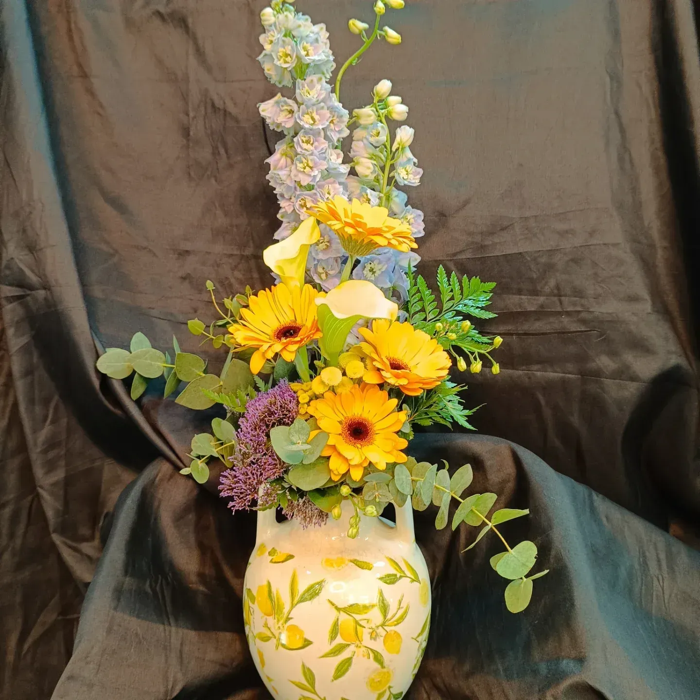 Arreglo floral en jarrón decorado con limones, gerberas amarillas, delfinios azules y vegetación.