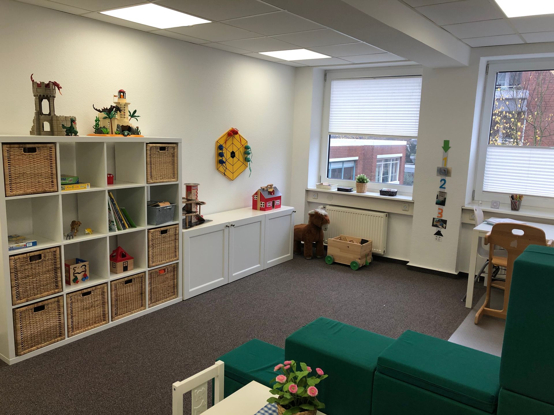 Spielzimmer mit weißen Regalen, Spielzeug, Fenstern und grünen Sitzmöbeln.