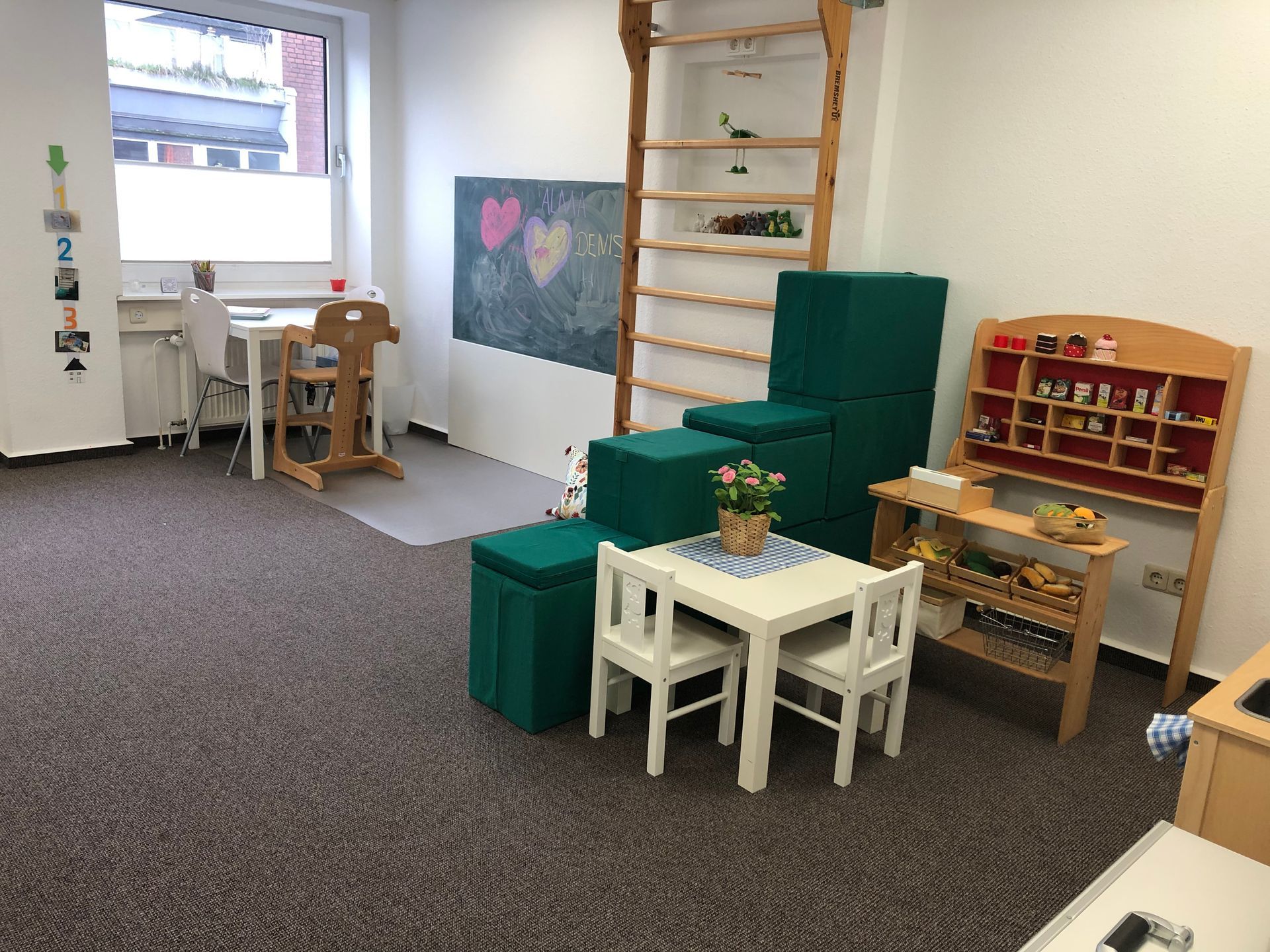 Spielzimmer für Kinder mit kleinem Tisch, Stühlen und Regalen an einer Wand mit Tafel, Leiter und Fenster.