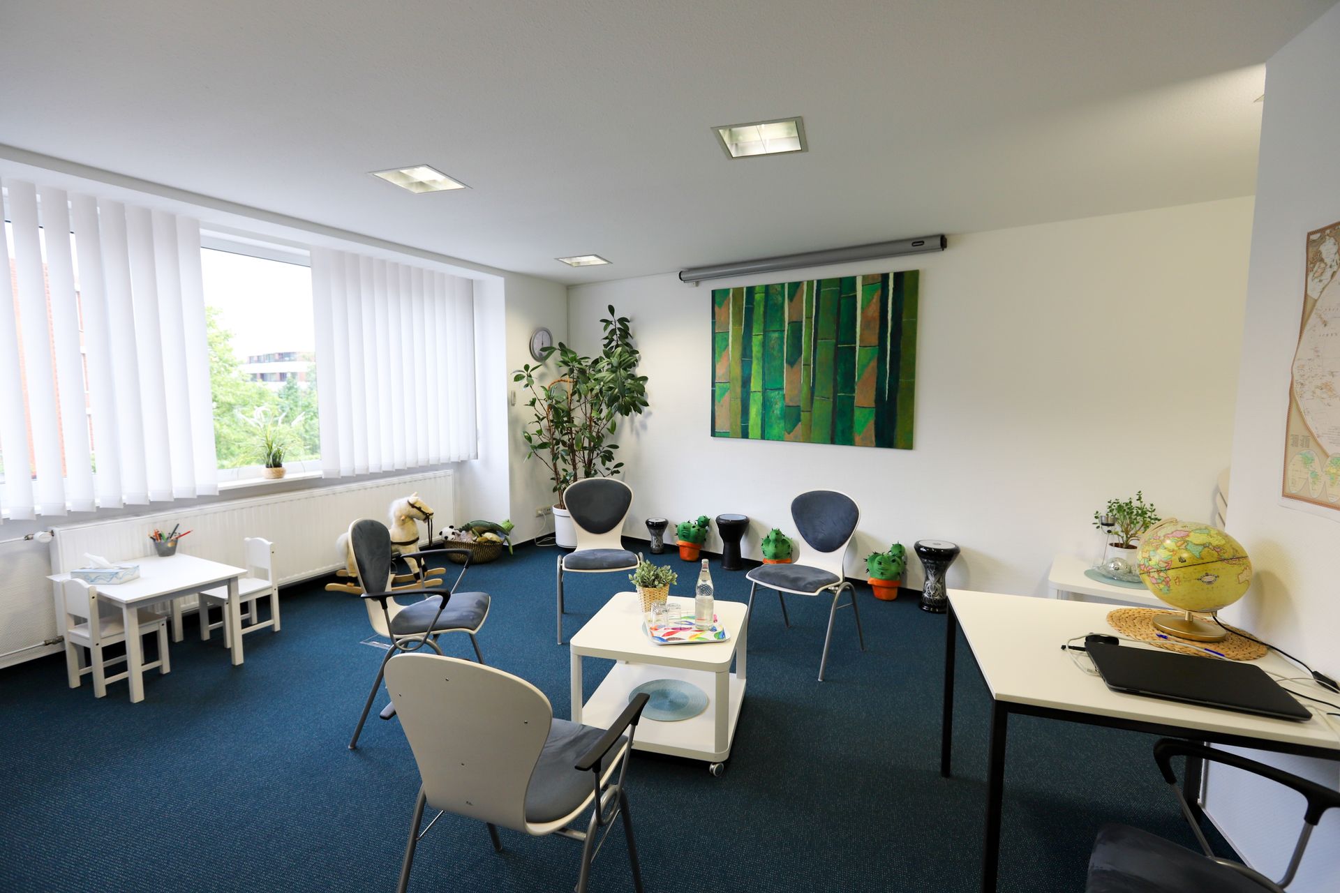 Therapieraum mit Stühlen, kleinen Tischen, Pflanzen und farbenfrohen Kunstwerken auf blauem Teppich.