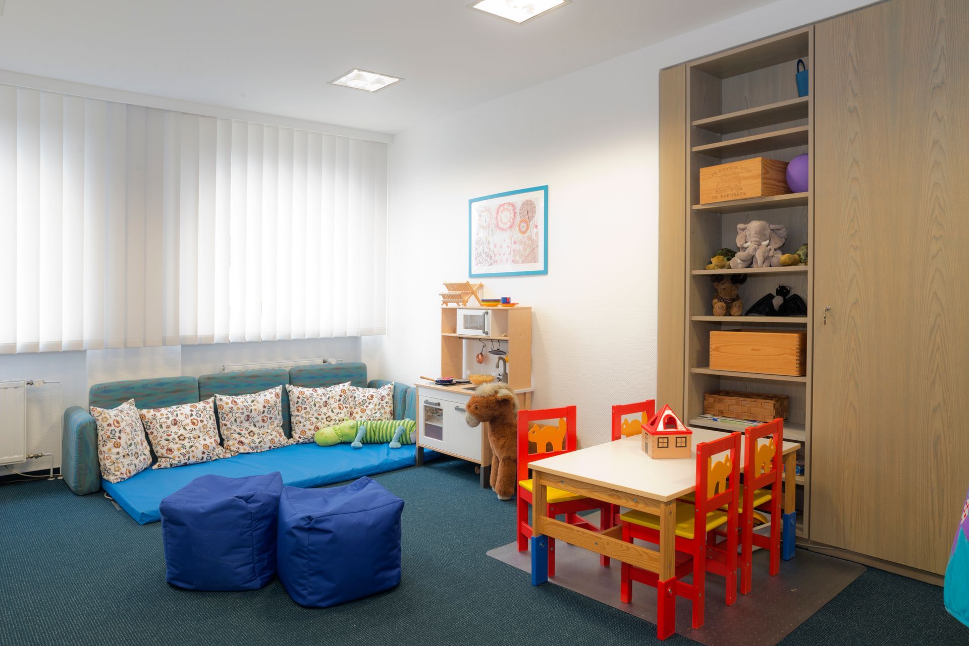 Spielzimmer mit blauem Sofa, Tisch mit roten Stühlen, Spielküche und Regal.