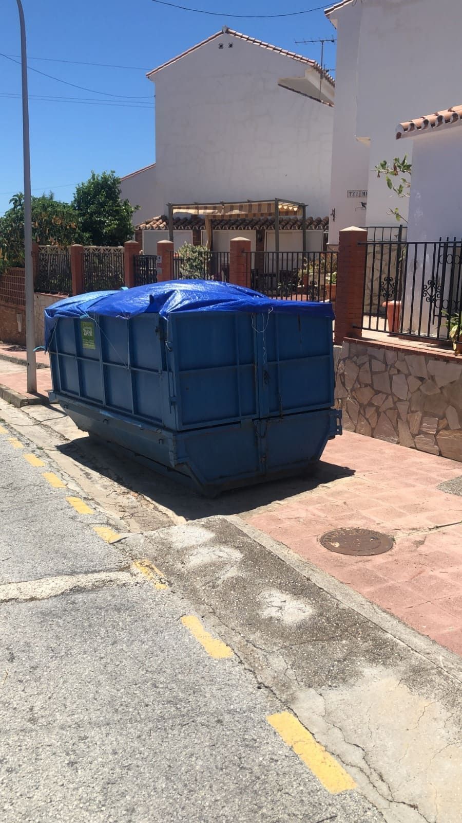Contenedor azul en una acera, cubierto con una lona, al lado de una carretera y casas en un día soleado.