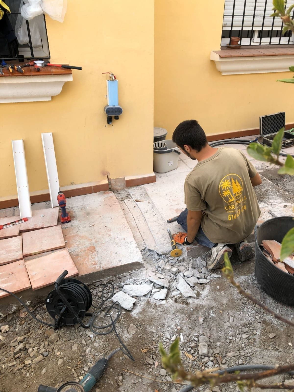 Hombre quitando baldosas del patio. Se ven una pared beige, escombros, herramientas y baldosas nuevas.