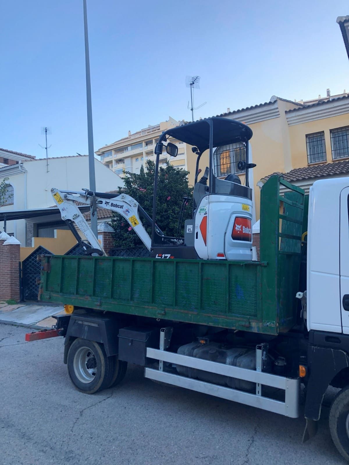 Excavadora Bobcat blanca en la plataforma de un camión volquete verde en una calle.