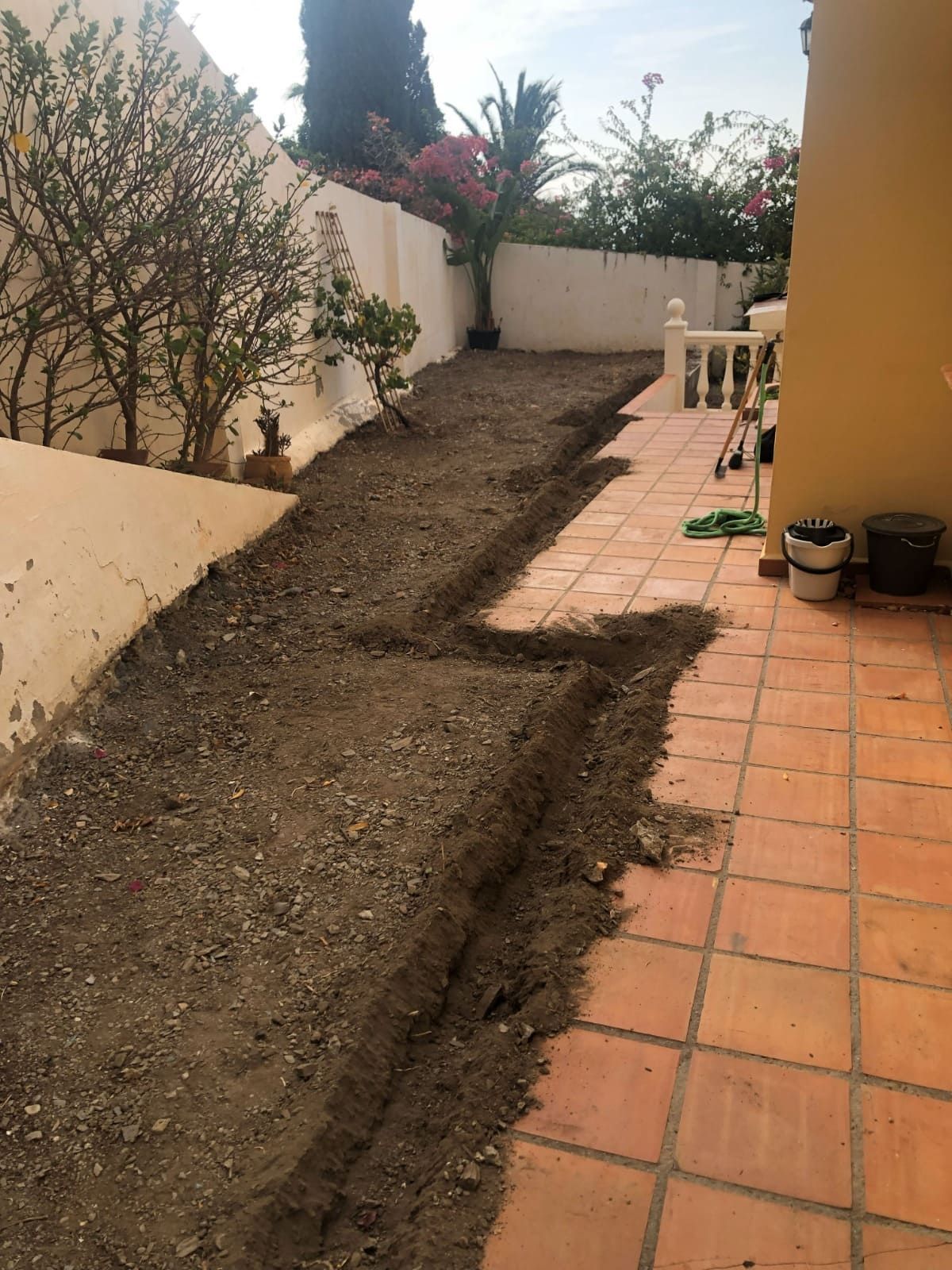 Un cantero parcialmente cavado junto a un patio de baldosas y paredes blancas; hay tierra presente.