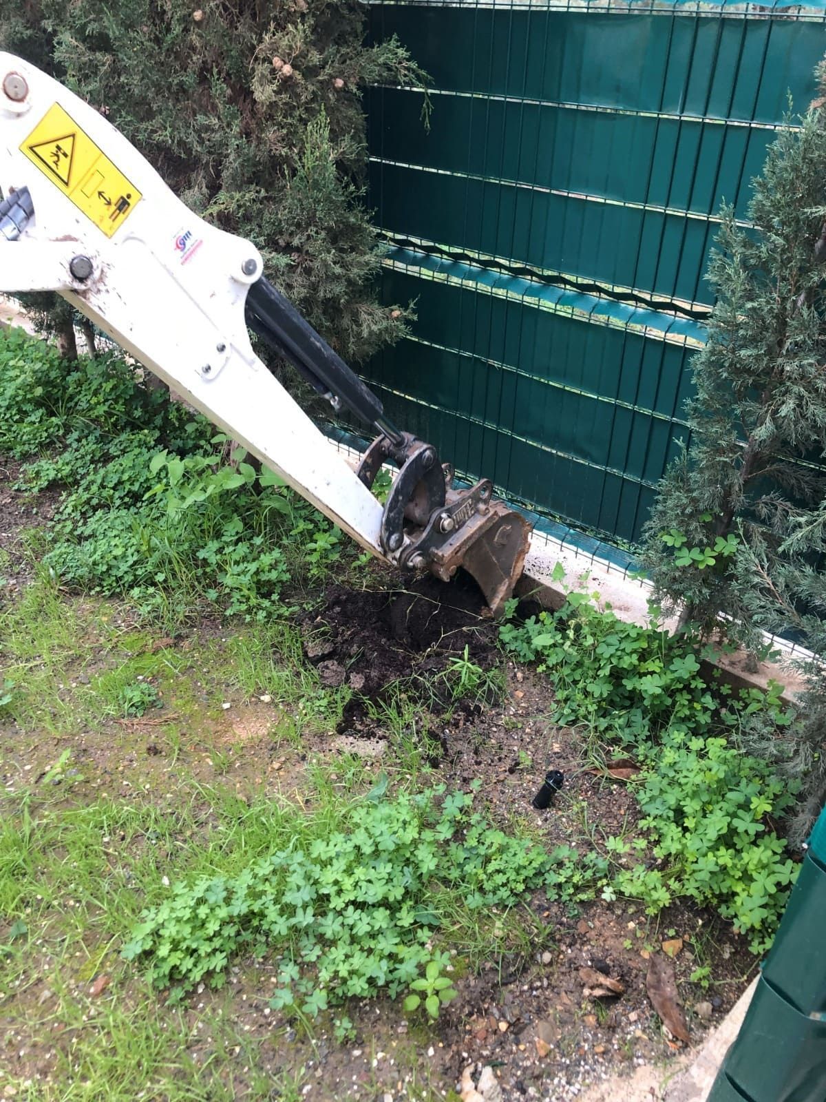 Una pequeña excavadora excava cerca de una valla verde y follaje.