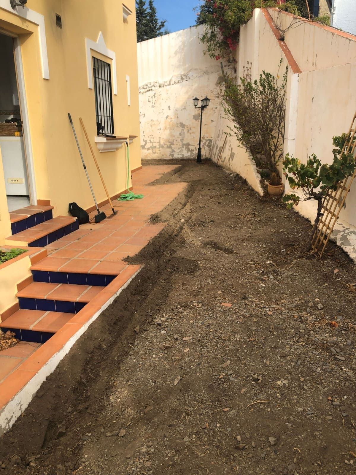 Un arriate estrecho y lleno de tierra junto a un patio de baldosas y una pared blanca. Hay herramientas en el patio.
