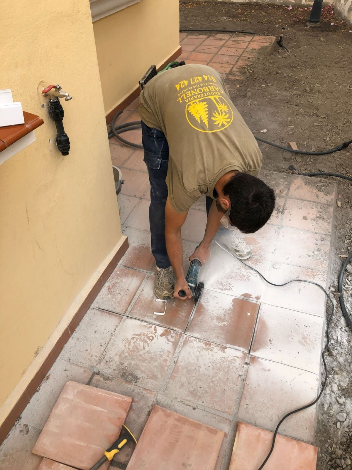 Una persona usa una amoladora para cortar baldosas en un patio. Lleva camisa de trabajo y vaqueros.