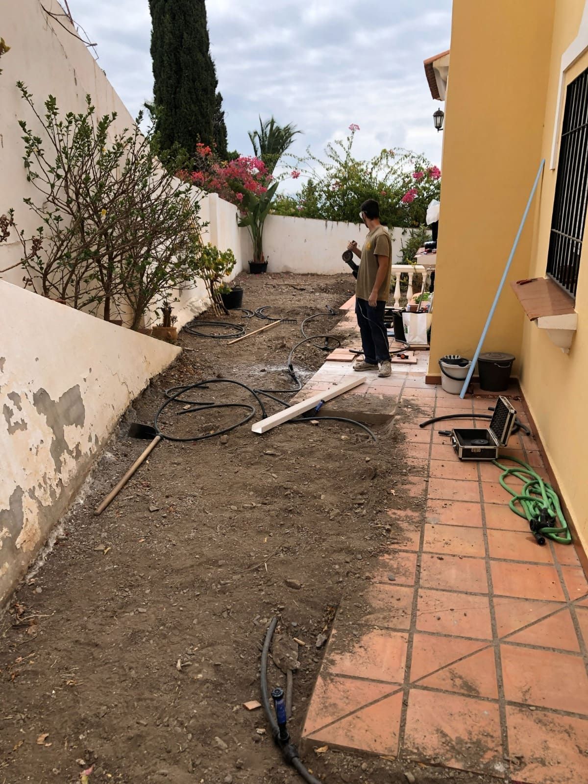 Una persona se encuentra en un patio lleno de tierra con herramientas de jardinería y una casa a cada lado bajo un cielo nublado.