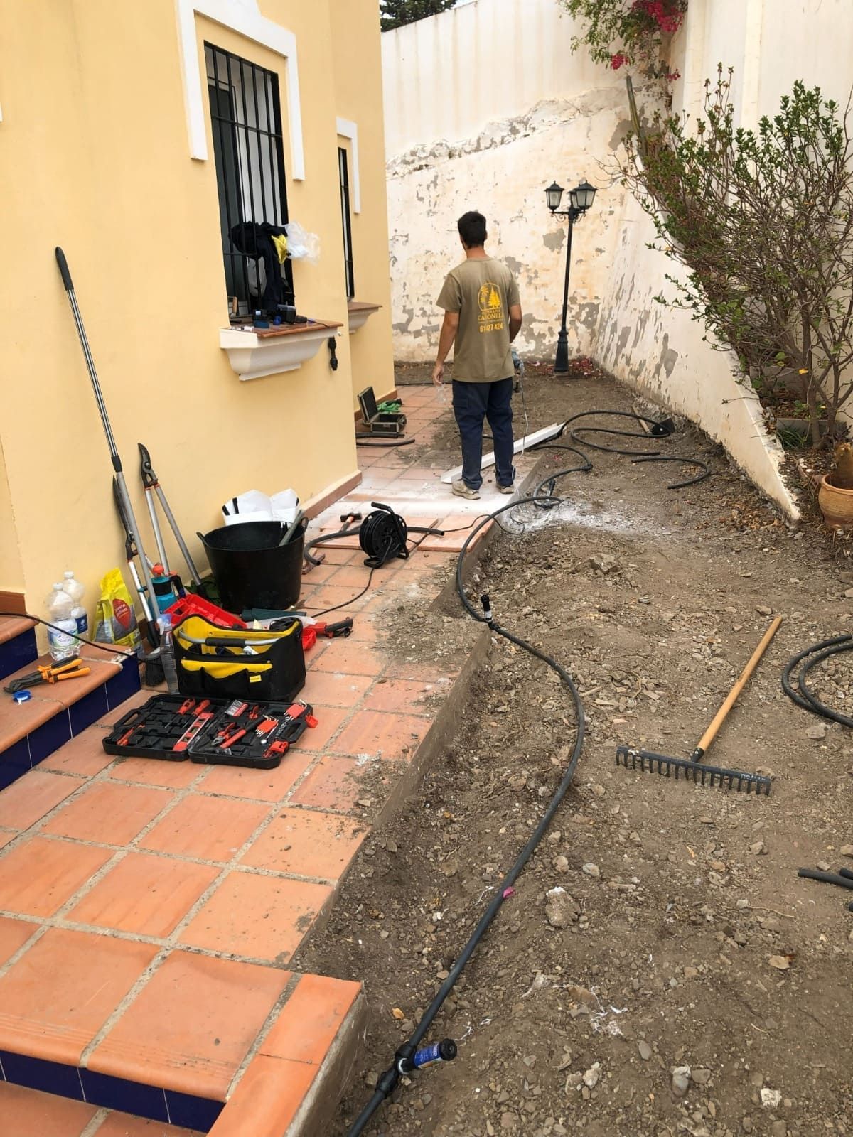 Hombre de pie junto a un patio con obras de riego en marcha. Se ven herramientas y suministros en el suelo.