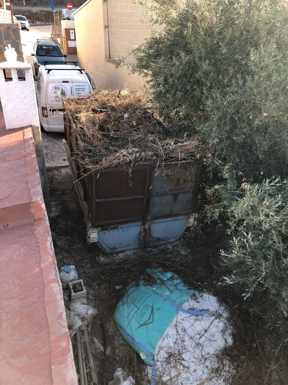 Un contenedor de basura marrón lleno de escombros y rodeado de basura, con una camioneta blanca y follaje cerca.