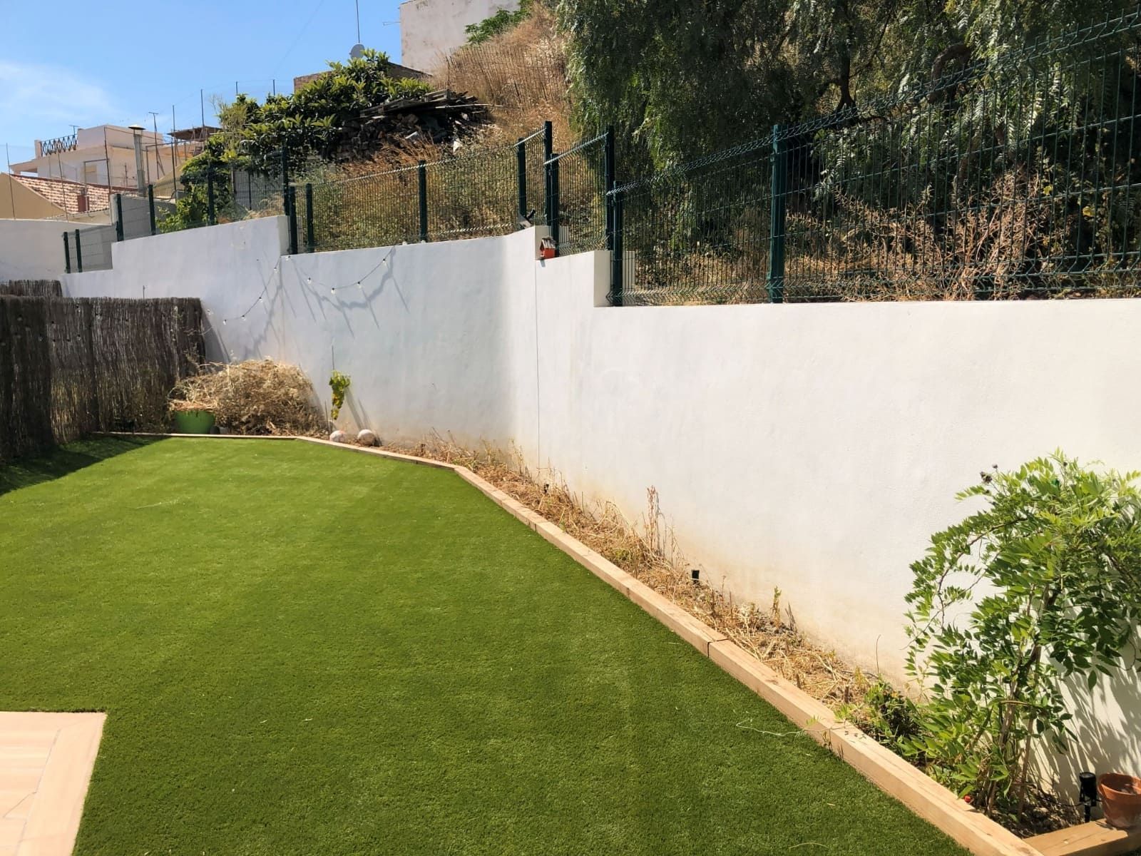 Césped artificial verde bordeado por una pared blanca y plantas secas; una valla remata la pared.
