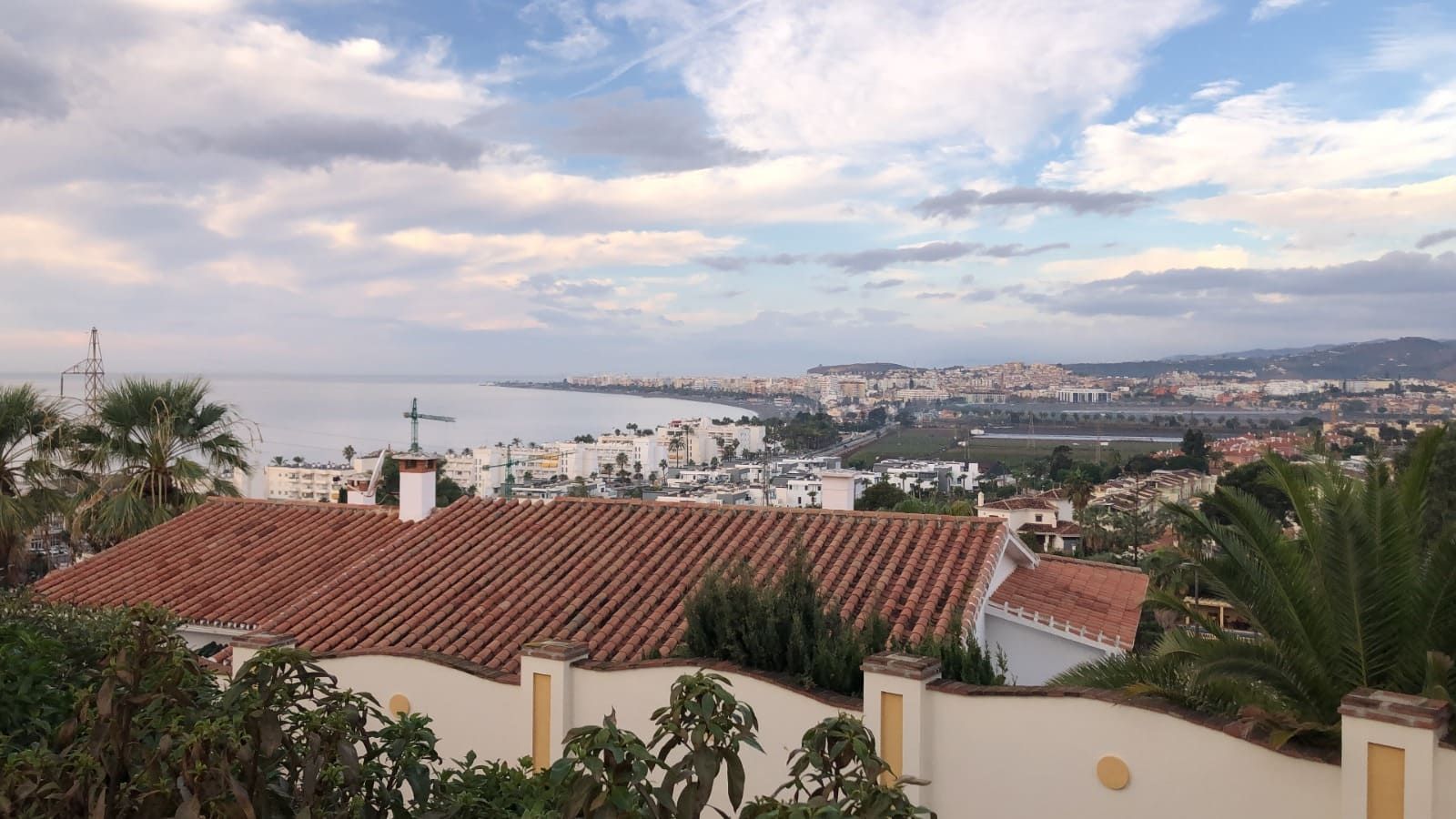 Vista desde la azotea de una ciudad costera, el mar y el cielo nublado. Tejados marrones, edificios blancos y palmeras verdes.