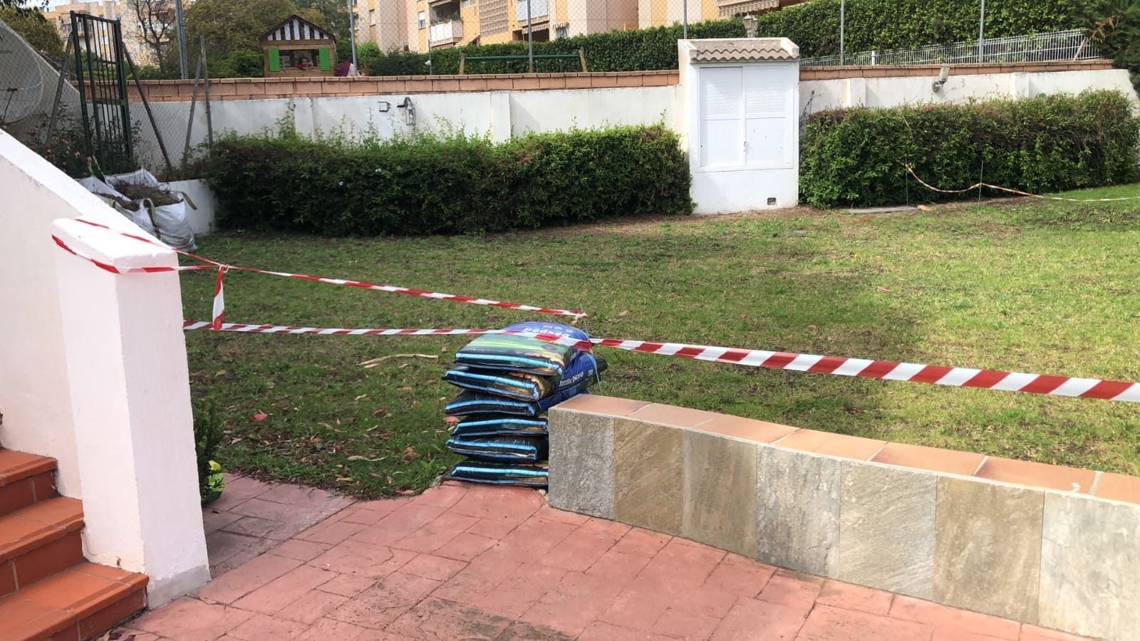 Césped con una pila de bolsas, delimitado por cinta de precaución y muro bajo, arbustos al fondo.