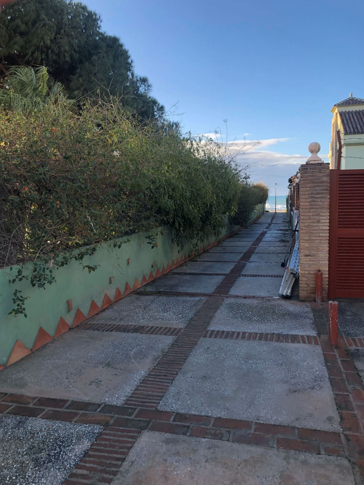 Camino de piedra bordeado de arbustos verdes y una pared de ladrillos, que conduce al mar bajo un cielo azul.