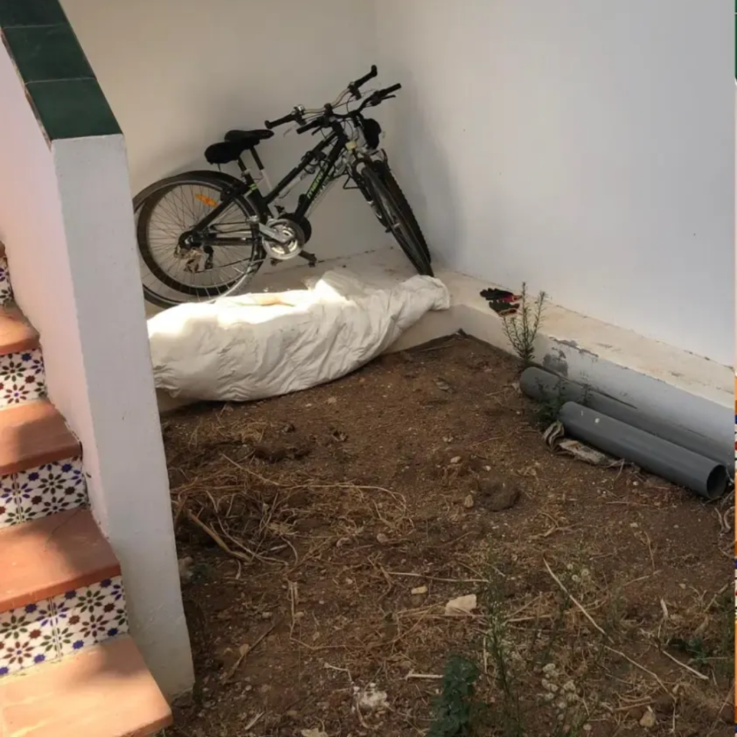 Una bicicleta negra está apoyada contra una pared blanca en una esquina. Suelo de tierra con tubos de plástico.