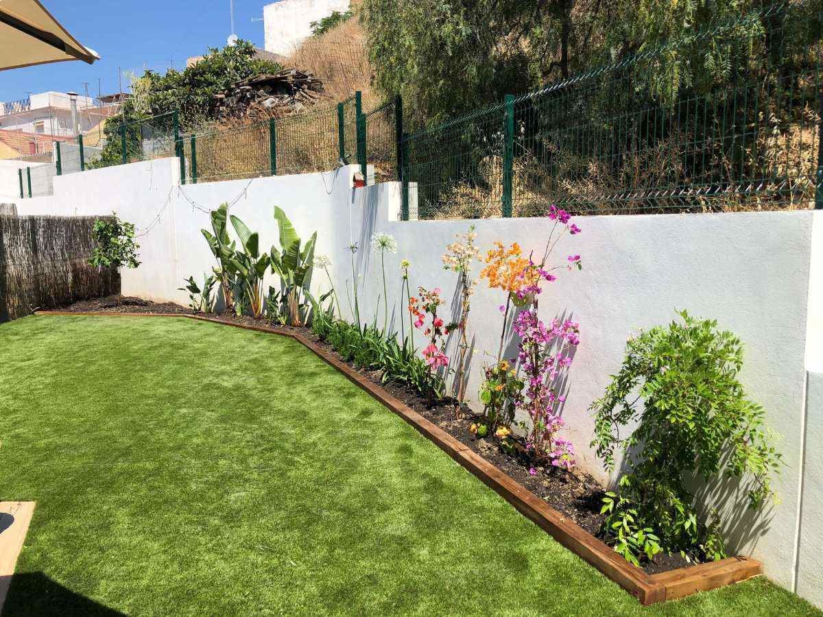 Patio trasero con césped artificial, parterre junto a una pared blanca y flores pintadas.
