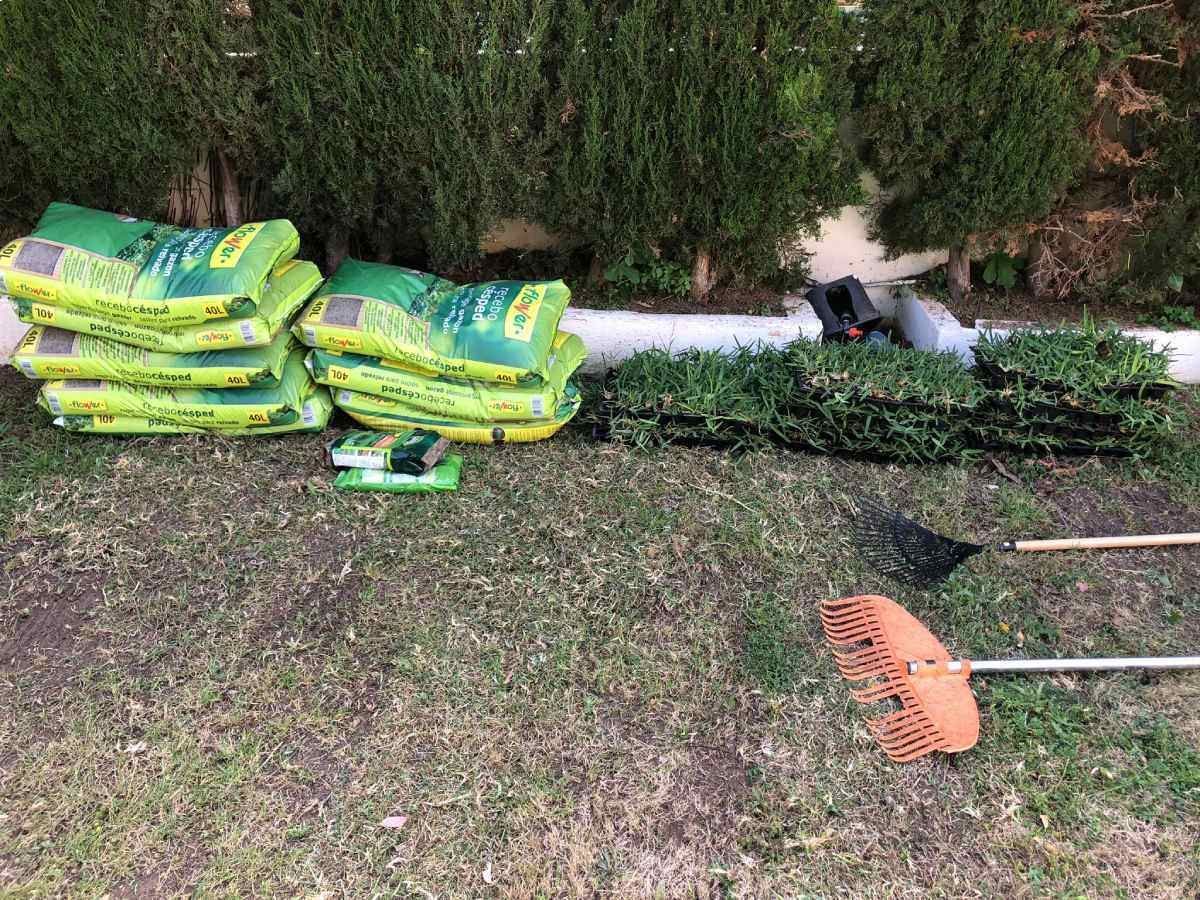 Bolsas verdes de tierra, setos recortados y herramientas de jardinería reposan sobre el césped en un patio.
