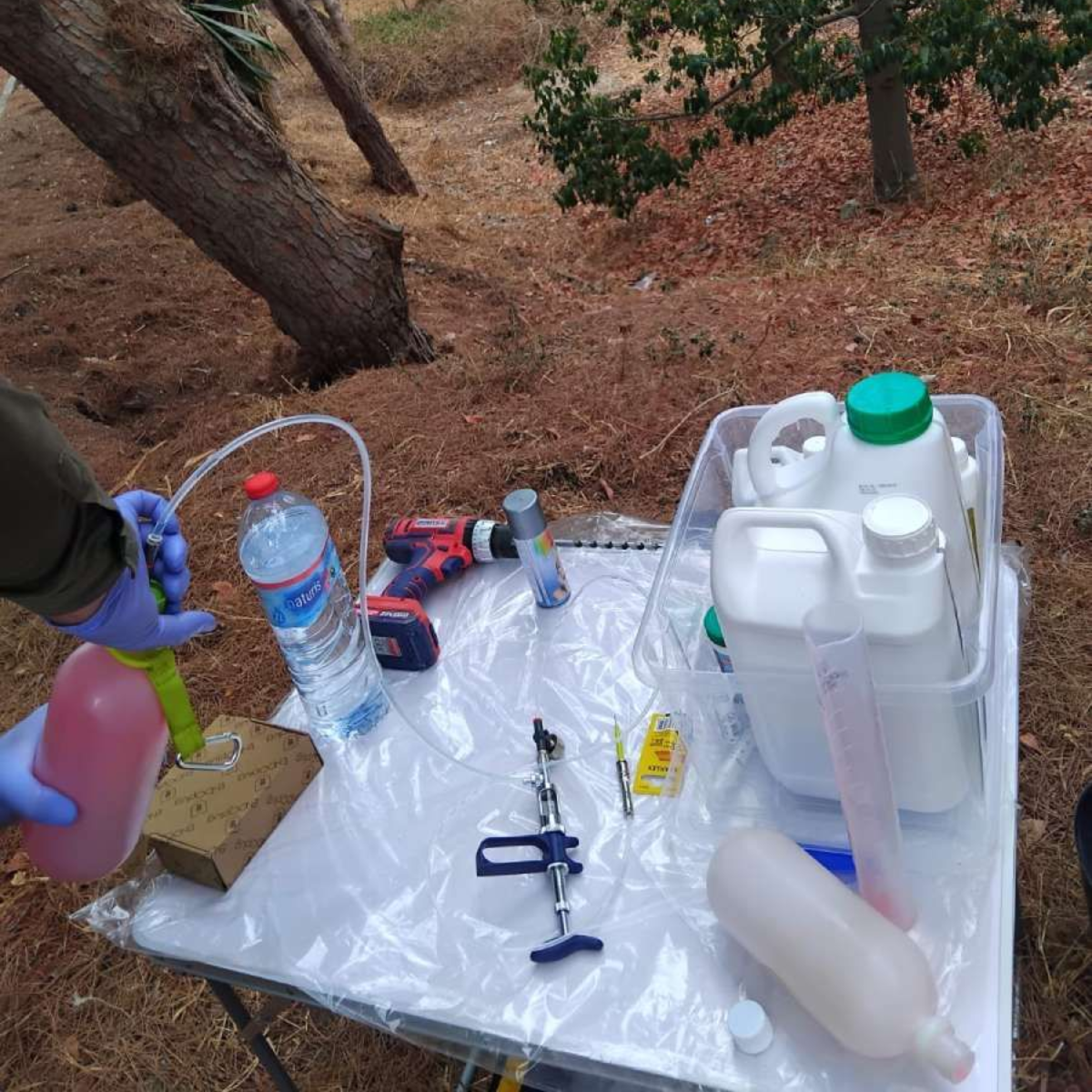 Una persona con guantes utiliza equipo sobre una mesa en un entorno exterior. Hay varias botellas, un taladro y tubos.