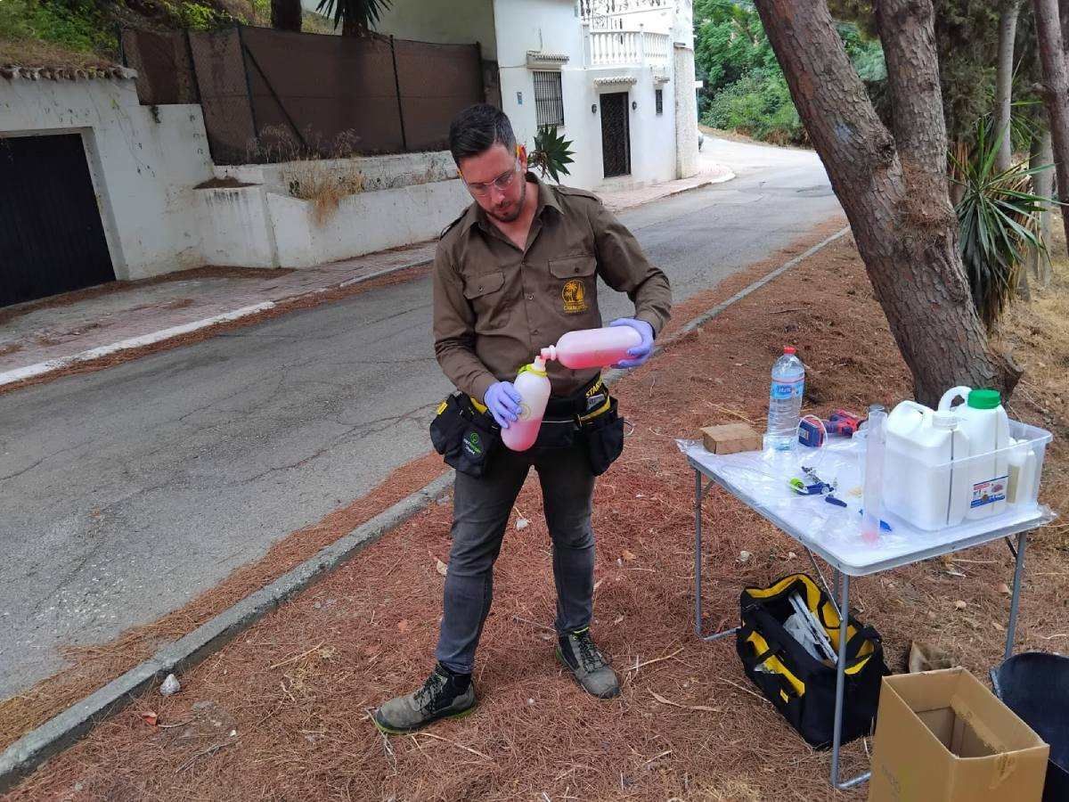 Un hombre uniformado vierte un líquido rosa de una botella al aire libre, cerca de una mesa con suministros.