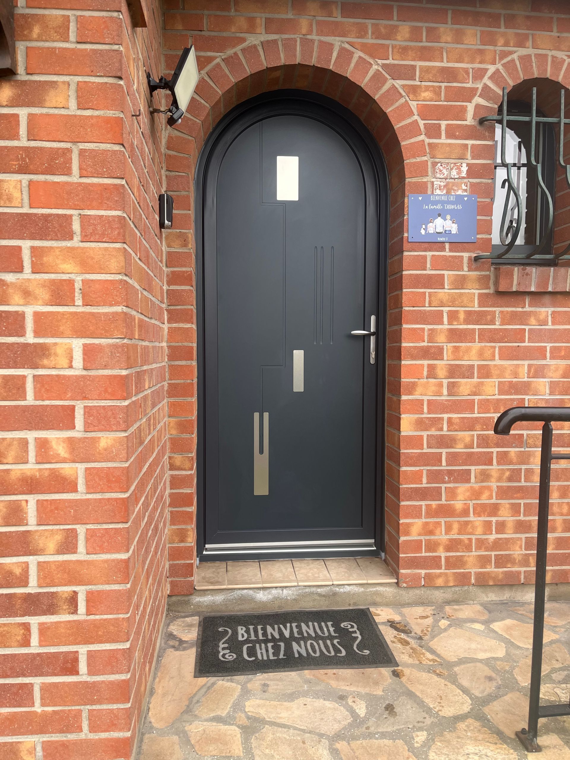 Porte d'entrée cintrée gris foncé avec rectangles décoratifs, extérieur en briques.