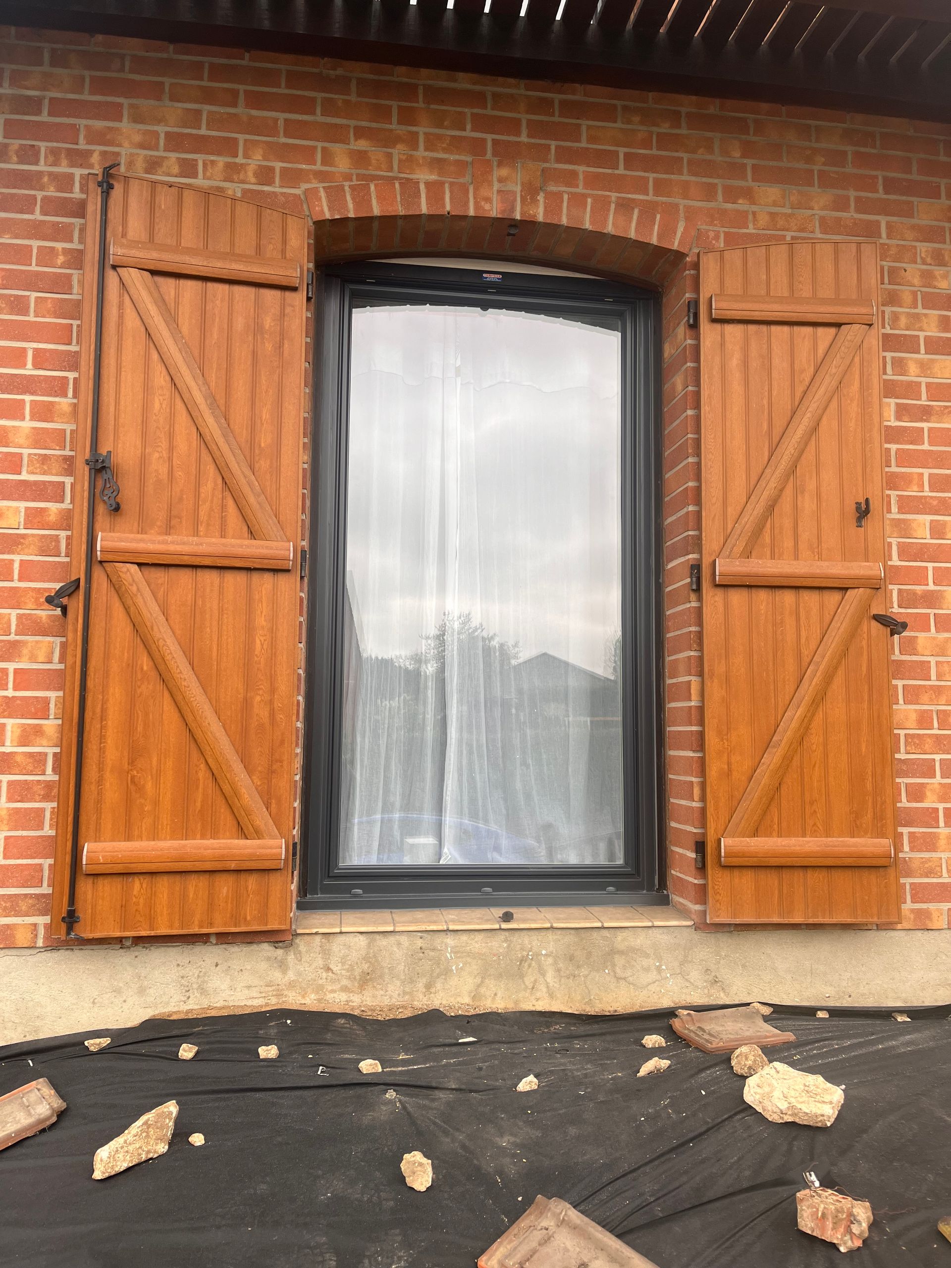 Porte-fenêtre à volets marron et cadre noir dans un mur de briques.