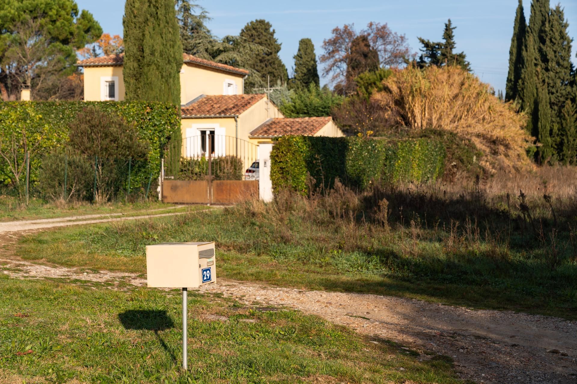 Maison rurale au toit de tuiles rouges, bordée de grands cyprès, avec une boîte aux lettres ensoleillée, le long d'un chemin herbeux.