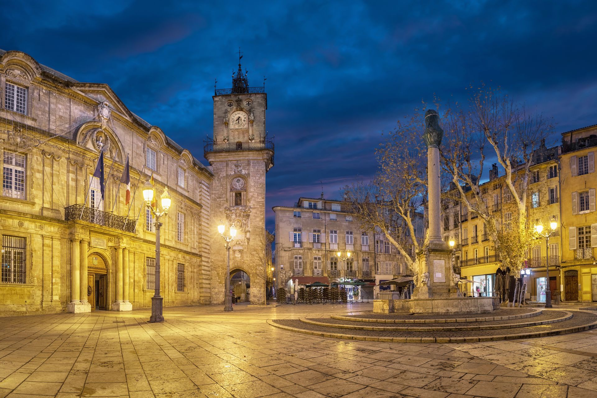 Place de l'Hôtel de ville à Aix-en-Provence illuminée la nuit, avec des bâtiments en pierre, une tour d'horloge et un pavage sous un ciel bleu.