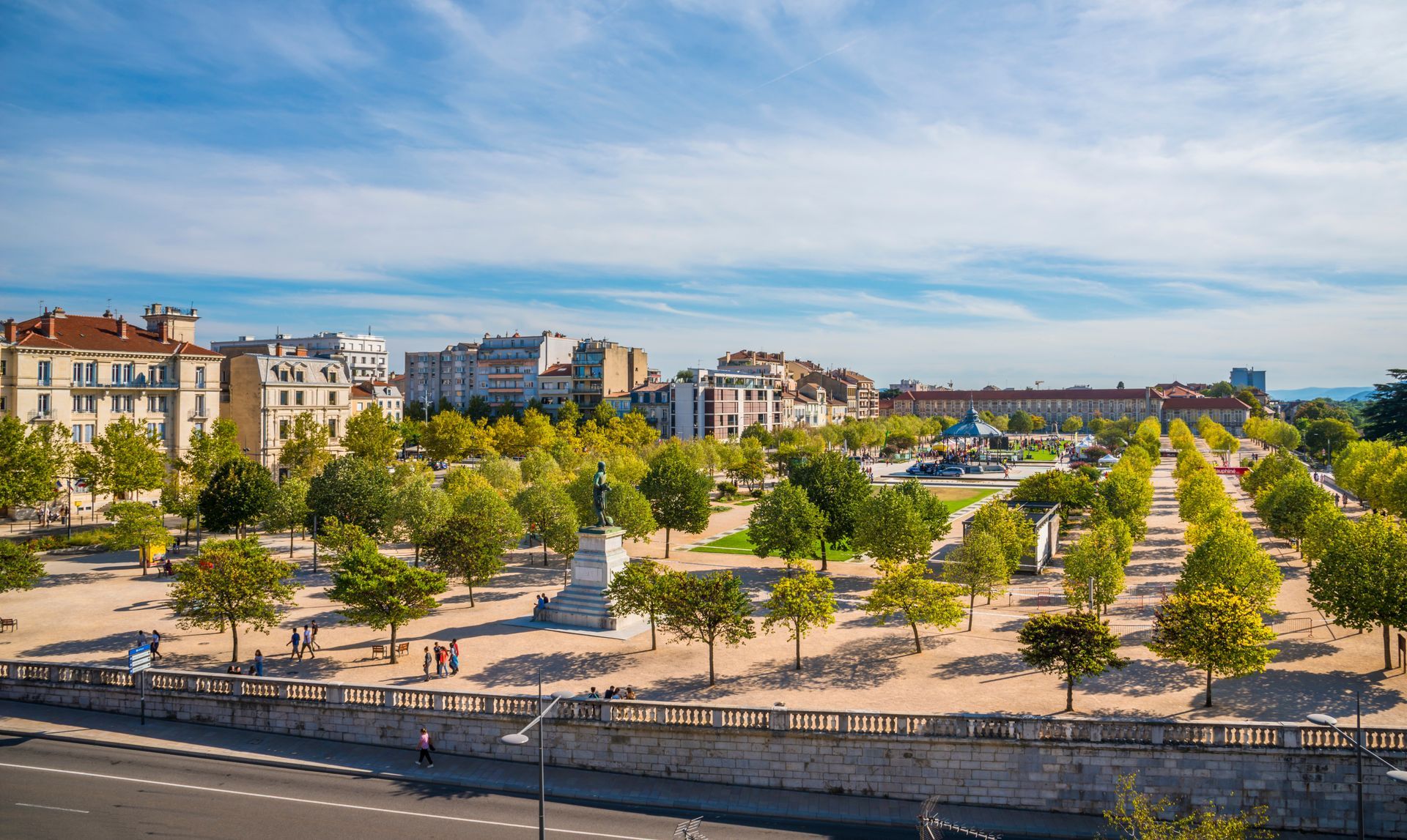 Vaste place à Valence, bordée d'arbres et de bancs, entourée d'immeubles d'habitation sous un ciel bleu