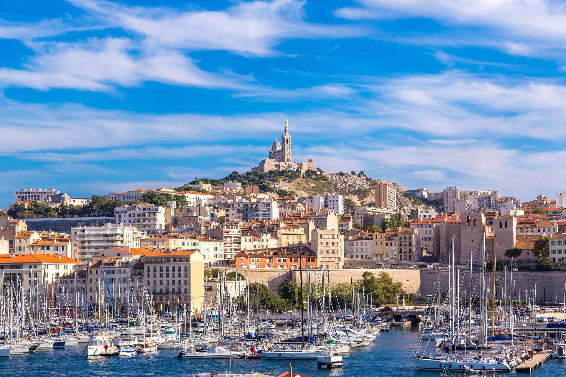 Le port de Marseille avec ses voiliers, ses bâtiments en bord de mer et sa basilique perchée sur une colline, sous un ciel bleu.