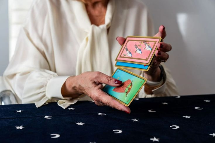 Des mains mélangeant des cartes de tarot colorées, roses, bleues et vertes, au-dessus d'un tissu foncé à motifs d'étoiles.