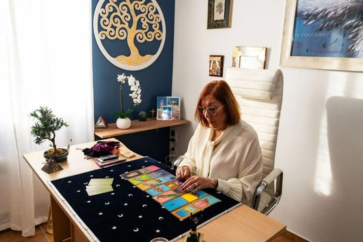 Une femme assise à une table dispose des cartes colorées dans une pièce ensoleillée ornée de tableaux.