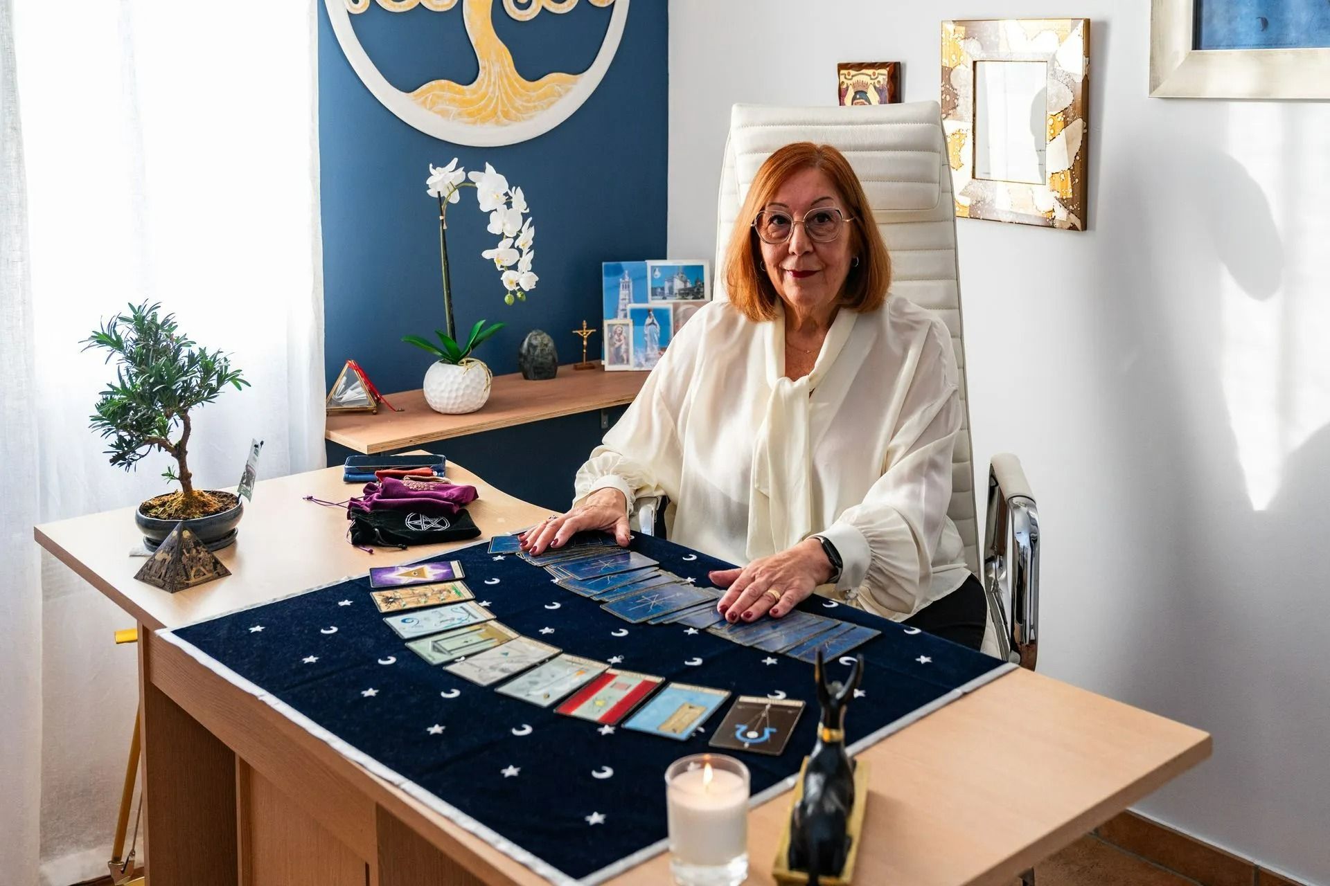 Une femme assise à une table avec des cartes de tarot dans une pièce lumineuse regarde la caméra.