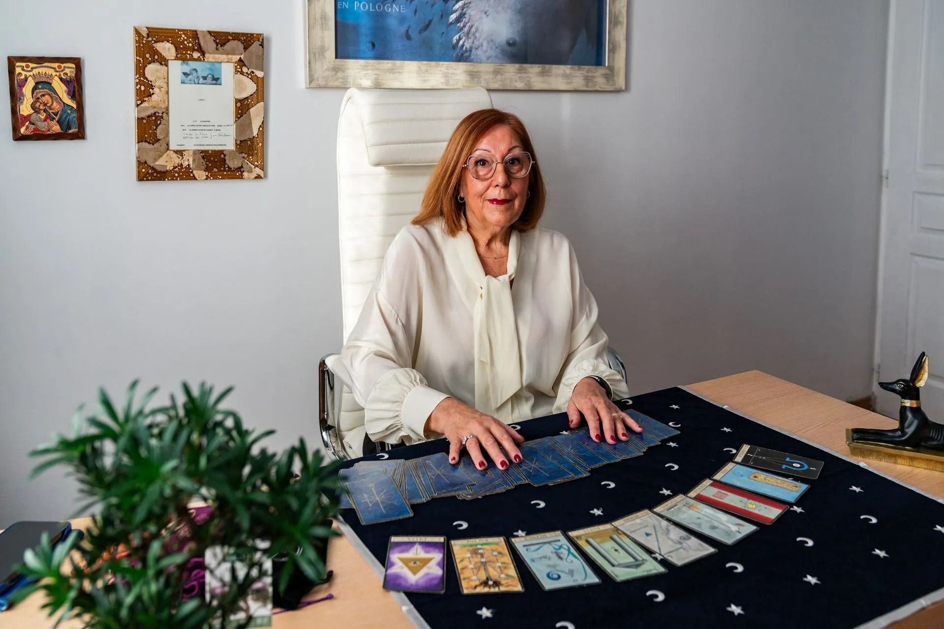 Une femme, assise à une table, dispose des cartes de tarot sur un tissu sombre dans une pièce lumineuse.
