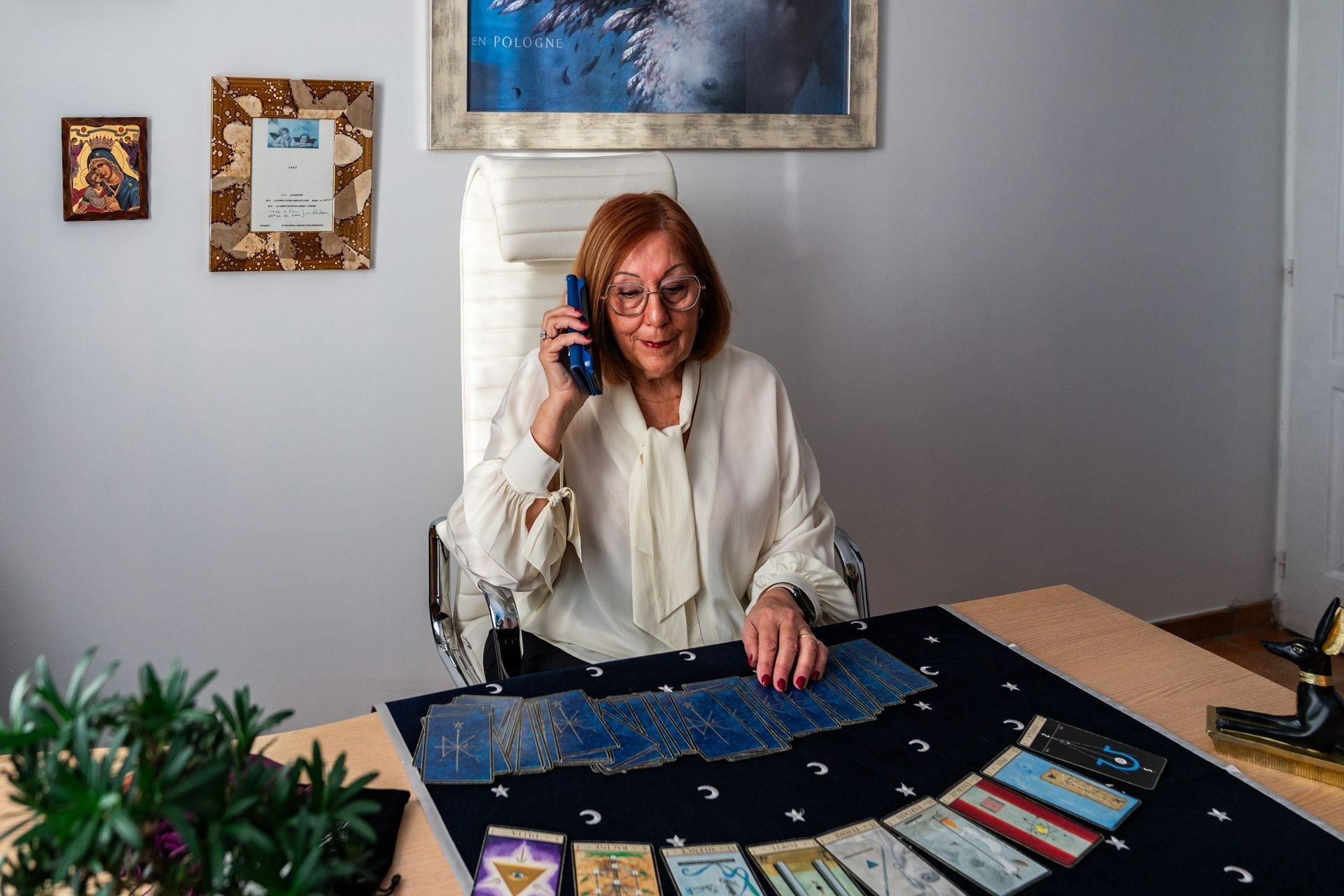 Une femme assise à une table, tenant un téléphone et regardant des cartes étalées dans une pièce lumineuse.