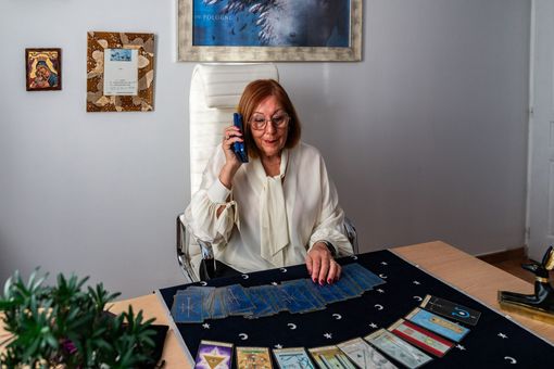 Une femme assise à une table, tenant des cartes et un téléphone, avec des cartes de tarot disposées dans une pièce.