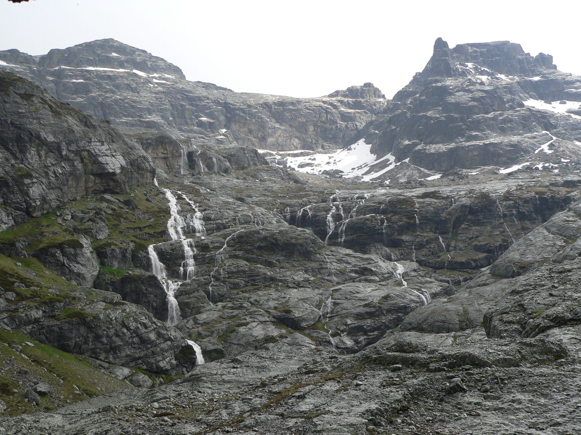 Paisaje de montaña rocosa con cascadas y parches de nieve.
