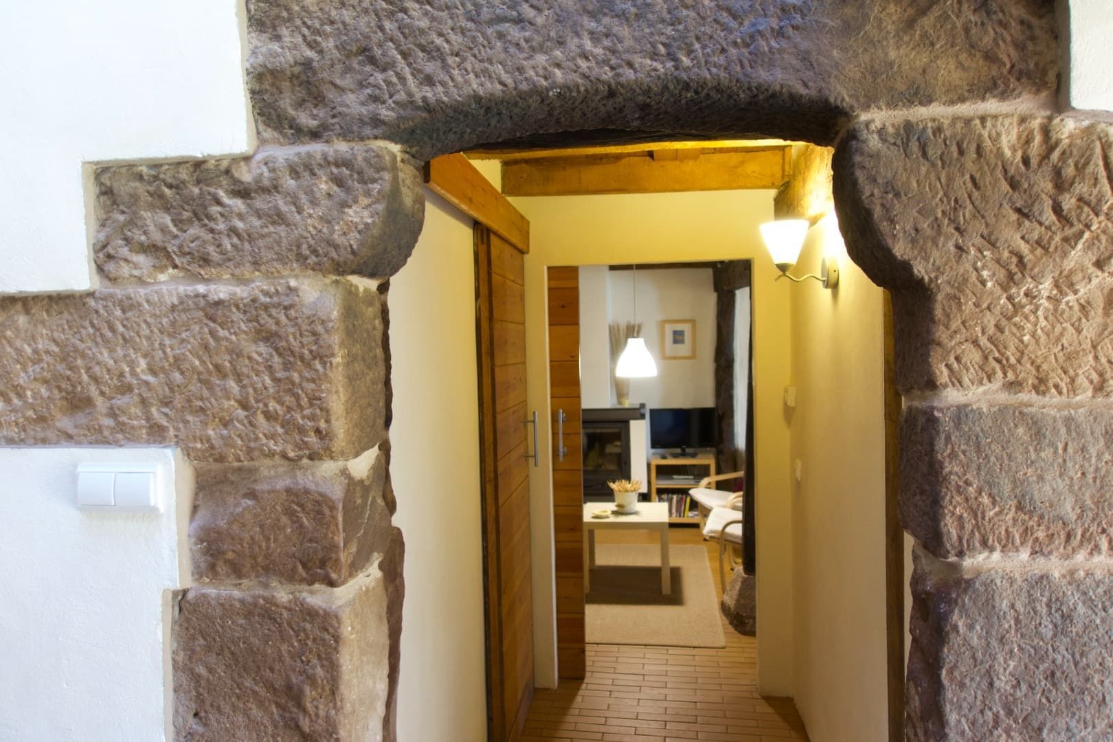 Una puerta de piedra conduce a un pasillo con puerta de madera, iluminado por apliques de pared.