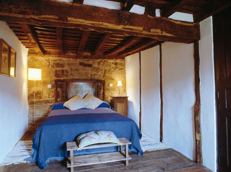 Dormitorio con vigas vistas, paredes de piedra y una colcha azul. Dos lámparas flanquean el cabecero.