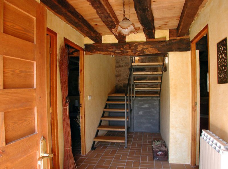 Pasillo con vigas de madera, escaleras y puertas abiertas. Tonos marrones, tostados y grises en todo el espacio.