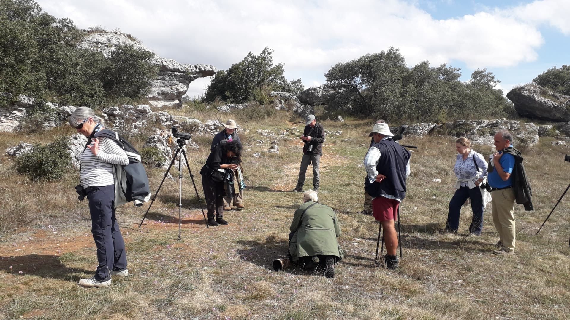 Grupo de personas con equipo, observando un arco natural en un campo de hierba, bajo un cielo soleado.