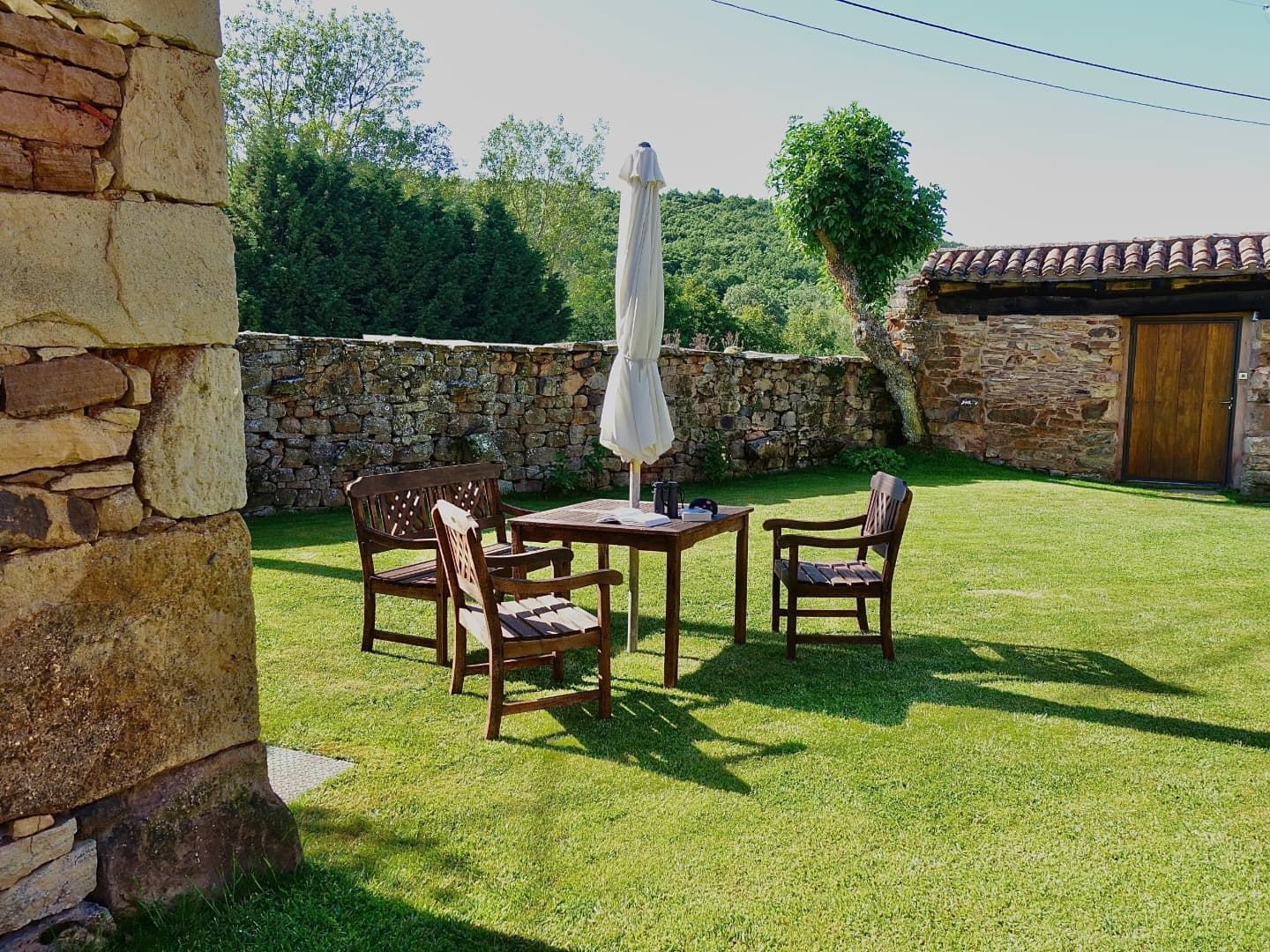 Patio al aire libre con mesa, sillas y sombrilla sobre césped verde, fondo de pared de piedra.