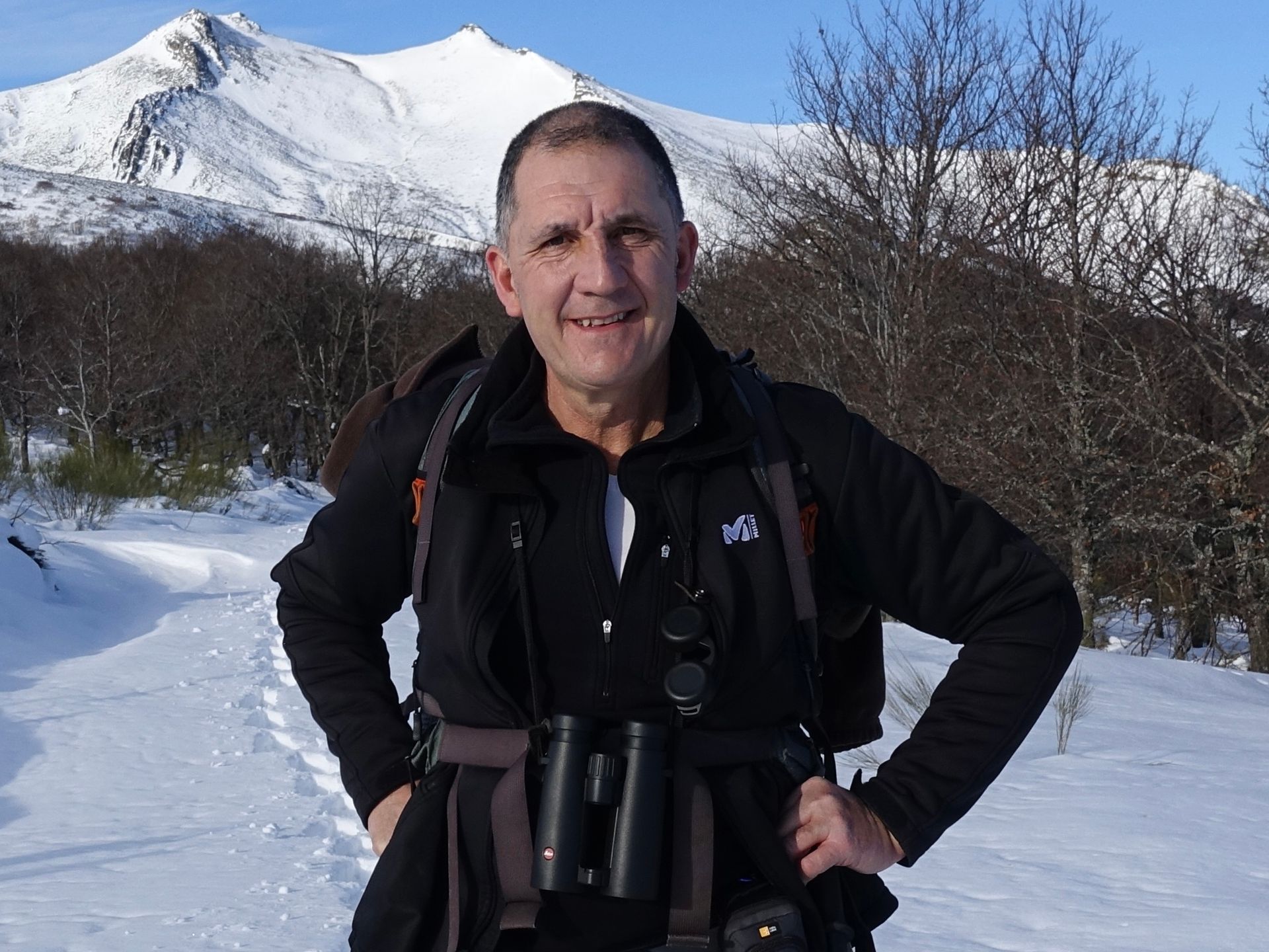 Un hombre con chaqueta negra se encuentra en un camino nevado con un fondo de montañas nevadas, sonriendo y sosteniendo binoculares.