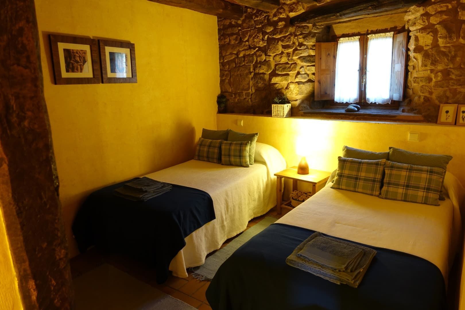 Dos camas individuales en una habitación rústica con pared de piedra, paredes amarillas, pequeña ventana y mesita de noche con lámpara.
